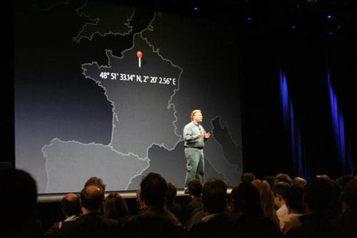 A speaker on stage in front of a large map projection, displaying geographic coordinates. The audience is seated in the foreground, attentively watching the presentation.
