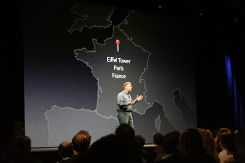 A speaker stands on stage presenting in front of a large map projection showing the location of the Eiffel Tower in Paris, France. An audience watches attentively in the foreground.