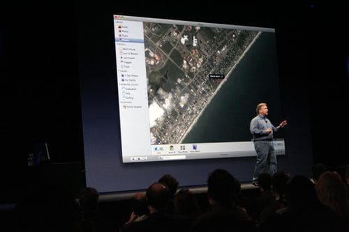 A man gesturing while presenting on stage, with a large screen displaying a satellite image of a coastal area. The audience, seated in the foreground, is attentively watching the presentation.