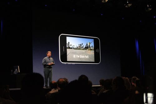 A speaker on stage presenting to an audience at a tech event, with a large smartphone graphic on the screen displaying a photo of a park. The setting has dim lighting, highlighting the speaker and the screen.