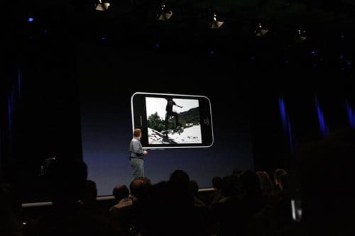 A presenter standing on stage in a well-lit auditorium, showcasing a large image of a person skiing on a smartphone screen. The audience is seated, with focus on the speaker and the displayed image.
