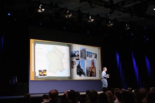 A presenter gestures while speaking on stage, with a large open presentation book on display. The book features a map, photos, and text, highlighting various locations. An audience watches attentively in a dimly lit auditorium.