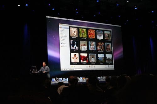 A speaker stands at a podium in front of a large screen displaying a photo management application. The screen showcases various images in a grid layout, framed by a dark auditorium with an audience visible in the foreground.
