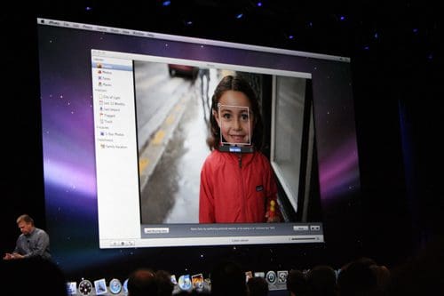 A screen displaying an image of a smiling girl wearing a red jacket with a background showing rain. The screen also shows photo editing software with facial recognition tools highlighted, while a presenter speaks in front of an audience.