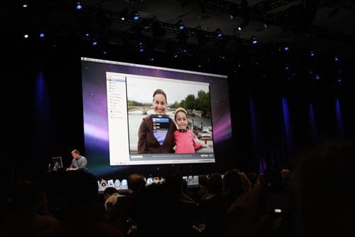 A large screen displaying a software demonstration during a conference, featuring a photo of two smiling individuals in a park. An audience is visible in the foreground, attentively watching the presentation.