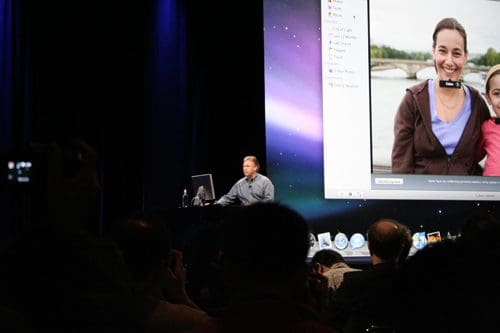 A speaker presenting at a tech event, standing behind a desk with a computer. The screen displays a photo of two smiling women outdoors. An audience is visible in the foreground, watching attentively.