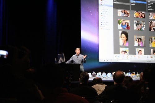 A speaker gesturing while presenting on stage, with a large display behind showing a photo management software interface. Audience members are visible in the foreground, attentively watching.