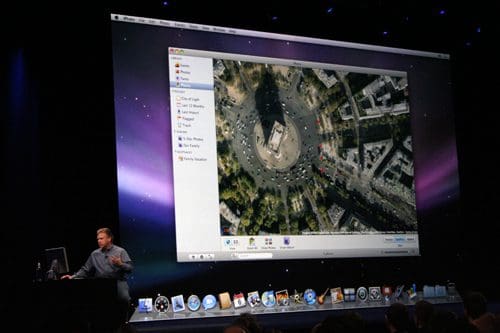 A man gives a presentation on stage with a large screen behind him displaying an aerial view of a city intersection, highlighting streets and buildings. The focus is on technology and urban landscape.