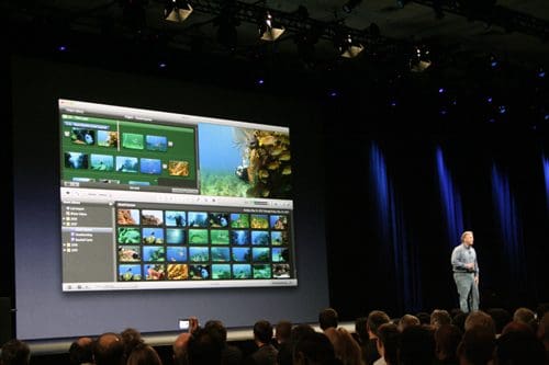 A speaker stands on a stage presenting to an audience, with a large screen displaying an editing software interface for images and videos featuring colorful underwater scenes and a library view.