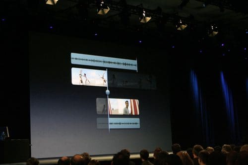 A large screen displaying a multimedia presentation featuring video clips and audio waveforms. An audience is seated in front, with a focus on the stage illuminated by spotlights.