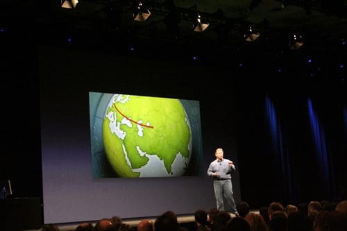 A speaker standing on stage with a presentation illustrating a green globe, showing a red line tracing a route across continents. The audience is seated in the foreground, attentively watching.