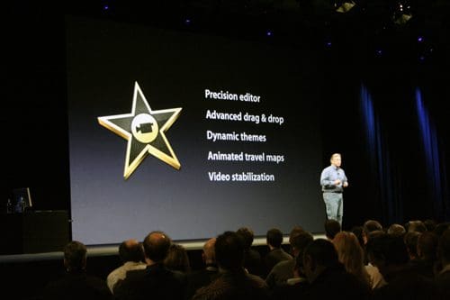 A speaker presenting on stage during a tech event, with a large screen showing features like 'Precision editor', 'Advanced drag & drop', and 'Video stabilization', while an audience attentively watches.