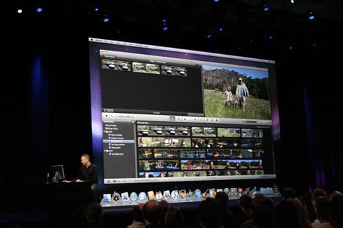 A presenter speaking on a stage with a large screen behind him showing video editing software, featuring a timeline and clips of outdoor scenes. An audience is seated in the foreground, engaged with the demonstration.