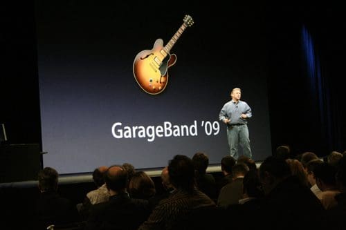 A speaker stands on stage, presenting at a tech event, with a large screen behind displaying the 'GarageBand '09' logo and an image of an electric guitar. An audience is visible in the foreground, focusing on the presentation.