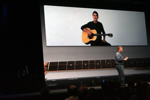 A speaker stands on stage while a large screen displays a young man playing an acoustic guitar. The backdrop features a guitar neck design, and the audience can be seen seated in darkened seats.