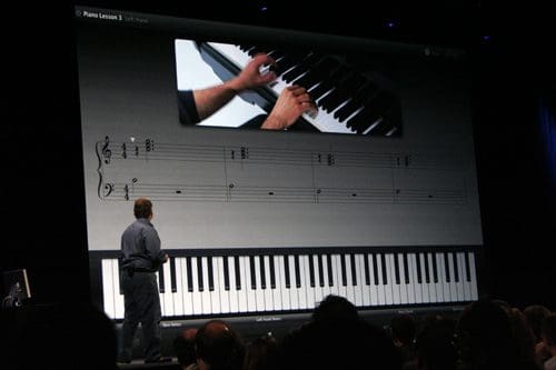 A presenter stands in front of a large screen displaying a piano lesson, featuring sheet music and a close-up of hands playing keys. An audience watches the demonstration attentively.