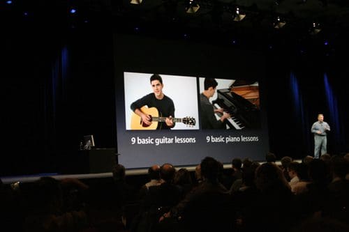 A speaker presenting on stage in a dimly lit auditorium, with a large screen displaying two men, one playing guitar and the other at a piano, promoting '9 basic guitar lessons' and '9 basic piano lessons.'