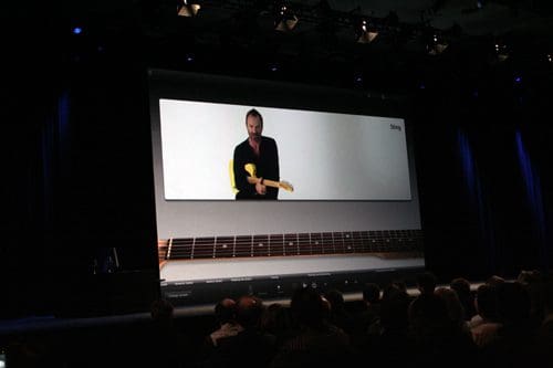 A man demonstrating guitar playing techniques on a large screen during a presentation. The stage is dimly lit with blue spotlights, and an audience is visible in the foreground.