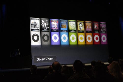 A display featuring multiple colorful iPod models lined up, showcasing various album covers on their screens. The setting appears to be a presentation, with an audience visible in the foreground.