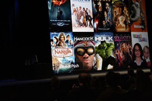 A large screen displaying movie posters, including titles like 'Narnia', 'Hancock', 'Hulk', and 'Sex and the City'. In the foreground, several silhouettes of people are seated, facing the screen.