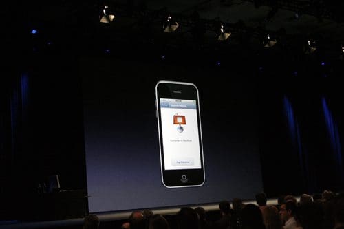 An iPhone displayed against a dark stage backdrop, showing a connection screen to a MacBook. Audience members are visible in the foreground, watching the presentation with interest.