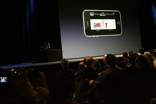 A crowd of attendees seated in a dimly lit auditorium, watching a presentation featuring a smartphone displaying images on its screen. A black table with a computer is visible in the background.