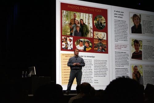 A speaker stands on stage in front of a large display showcasing family photos and text highlights from the year. The background includes colorful images of people in nature and written reflections.