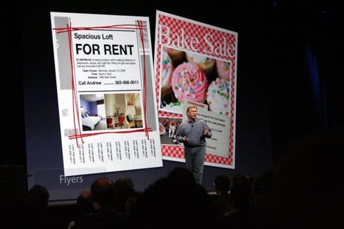A speaker standing on stage in front of a large screen displaying two flyers: one for a spacious loft for rent with a photo of a living room, and another promoting a bake sale with colorful cupcakes.