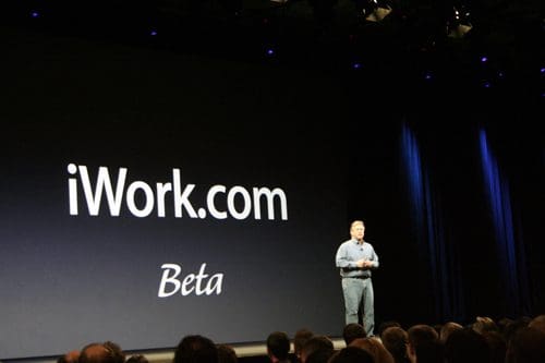 A speaker stands on stage in front of a large screen displaying "iWork.com Beta". The dark background contrasts with white text, while an audience sits below, attentively watching the presentation.