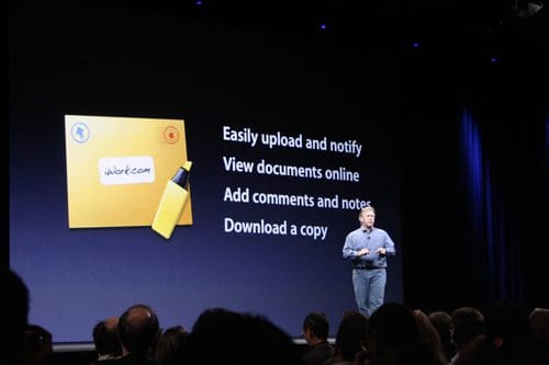 A speaker on stage presenting at a conference, standing in front of a large screen displaying features of iWork.com, including options to upload documents, view them online, add comments, and download files, with an audience in the foreground.
