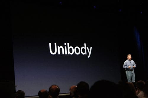 A presenter standing on stage in front of a large screen displaying the word 'Unibody' in bold white letters. The audience is partially visible in the foreground, seated and engaged.