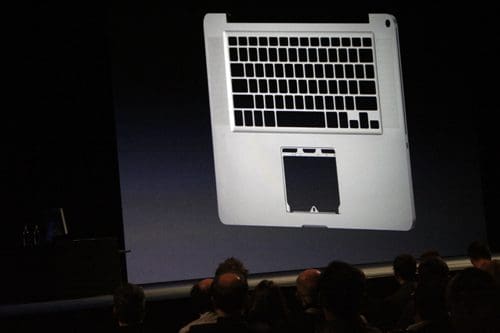 An overhead view of a laptop keyboard and trackpad displayed on a large screen during a presentation, with an audience visible in the foreground.
