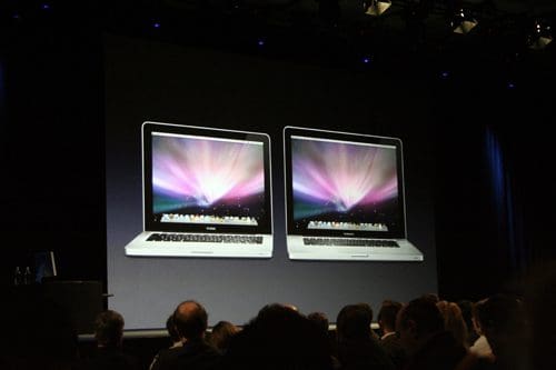 Two laptops displaying colorful screens with vibrant graphics, showcased on a large screen during a presentation, while an audience of people is seated below, focused on the display.