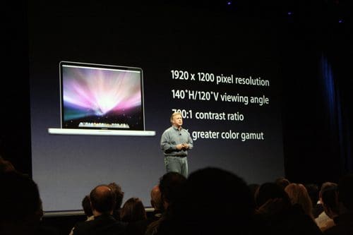 A speaker stands on stage presenting a laptop display, with specifications listed on a large screen behind him. The audience is seated, facing the presentation, under soft stage lighting.