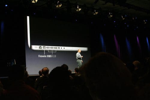 A speaker presenting on stage, pointing to a large screen displaying a laptop's connection ports, labeled "Firewire 800/2.0." The audience is visible in silhouette, attentively watching the presentation.