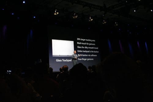 A presenter speaking at a conference, with a slide displaying features of a glass trackpad, including a larger tracking area and multi-touch gestures. The audience is partially visible in front of the stage.