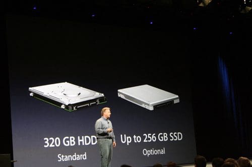 A speaker on stage presenting two types of storage options for a device, featuring a 320 GB hard disk drive (HDD) on the left labeled 'Standard' and a 256 GB solid-state drive (SSD) on the right labeled 'Optional.'