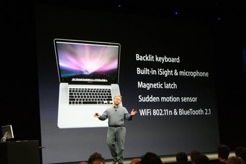 A presenter standing on stage with a laptop displayed on a large screen behind him. The screen shows features of the laptop, including 'Backlit keyboard', 'Built-in iSight & microphone', and 'WiFi 802.11n & Bluetooth 2.1'.