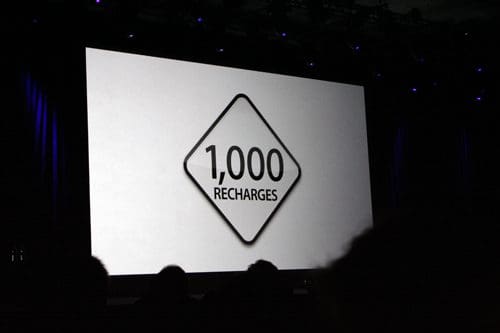 A large screen displaying the text "1,000 RECHARGES" inside a diamond shape, set against a plain white background. The stage is dimly lit with purple lighting, and silhouettes of an audience can be seen in the foreground.
