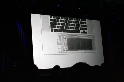 A projection on a screen displaying a schematic diagram of a laptop's internal components, including the keyboard at the top and battery layout beneath, with an audience silhouette in the foreground.