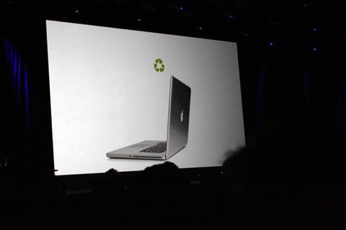 A large screen displaying a silver laptop shown slightly opened, with a recycling symbol above it. The background is dark with blue lighting accents, suggesting a tech presentation environment.
