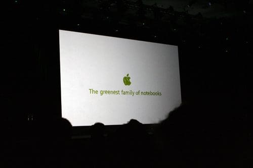 A dimly lit stage featuring a large screen displaying the Apple logo with the text "The greenest family of notebooks" in green font.