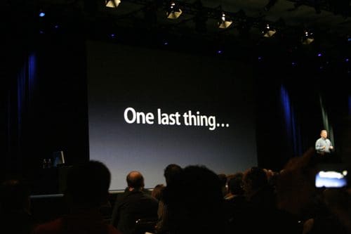 A large screen displaying the text "One last thing..." in bold white font, while a presenter stands in front of a seated audience in a dimly lit auditorium.