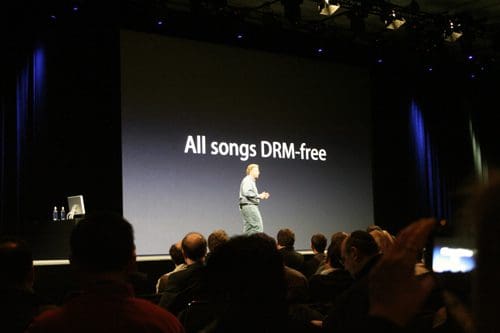 A presenter stands on stage in front of a large screen displaying the text "All songs DRM-free." The audience, seated in rows, watches attentively, with some individuals holding cameras.