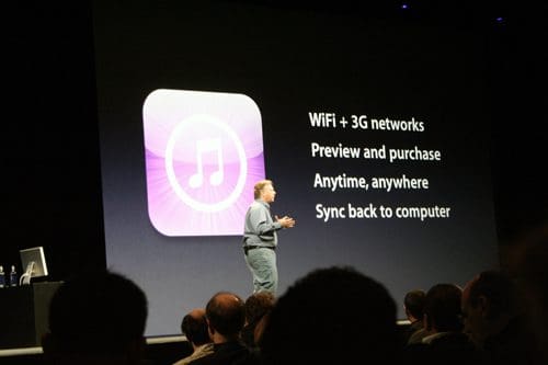 A speaker stands on stage in front of a large display showing an iTunes logo. Text beside the logo highlights features like WiFi and 3G networks, preview and purchase options, and syncing back to a computer. An audience is visible in the foreground.