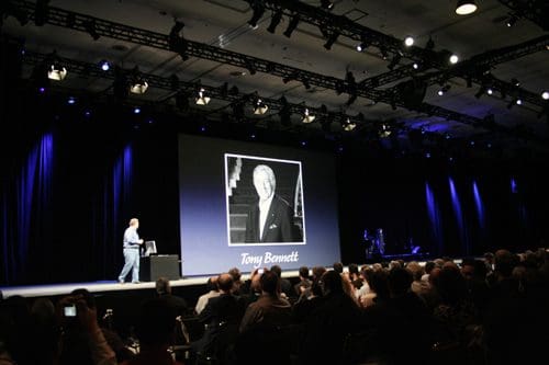 A speaker on stage presents to an audience in a large conference hall, with a projected photo of Tony Bennett on a screen behind. The hall is dimly lit with focused lighting on the stage.