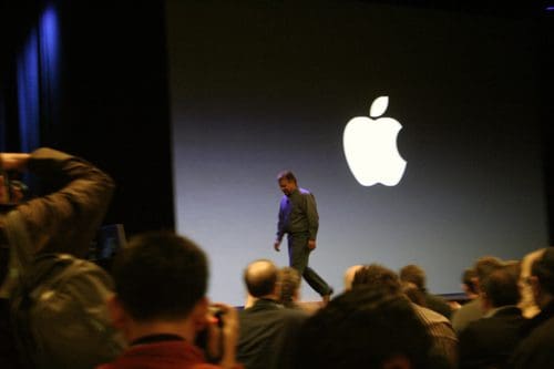 A speaker walking on stage in front of a large illuminated Apple logo. The audience is visible in the foreground, capturing the moment with cameras in a dimly lit venue.
