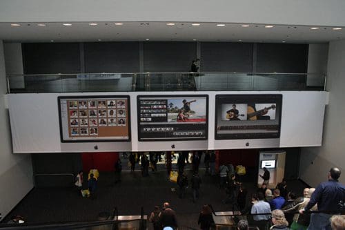 A large display wall inside a commercial space showcasing three digital screens featuring various images, including a grid of portraits, video editing software, and a musician playing guitar, while people walk by below.