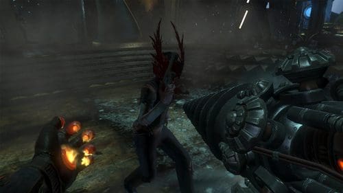 A first-person view of a character aiming a large drill weapon towards a humanoid figure with elongated, sharp features in a dimly lit environment. Fiery orange light flickers from the character's hand, contrasting the dark, atmospheric background.