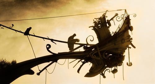 A silhouetted figure is seen working on an ornate, abstract sculpture resembling a ship's bow against a dramatic, cloudy sky, creating an artistic and serene atmosphere.